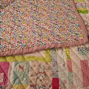 Könnte beinhalten: Eine Patchwork-Decke mit verschiedenen floralen und geometrischen Mustern in Rosa, Grün, Gelb und Blau. Die Decke zeigt ein kleines Bild einer Katze mit einer Schleife.