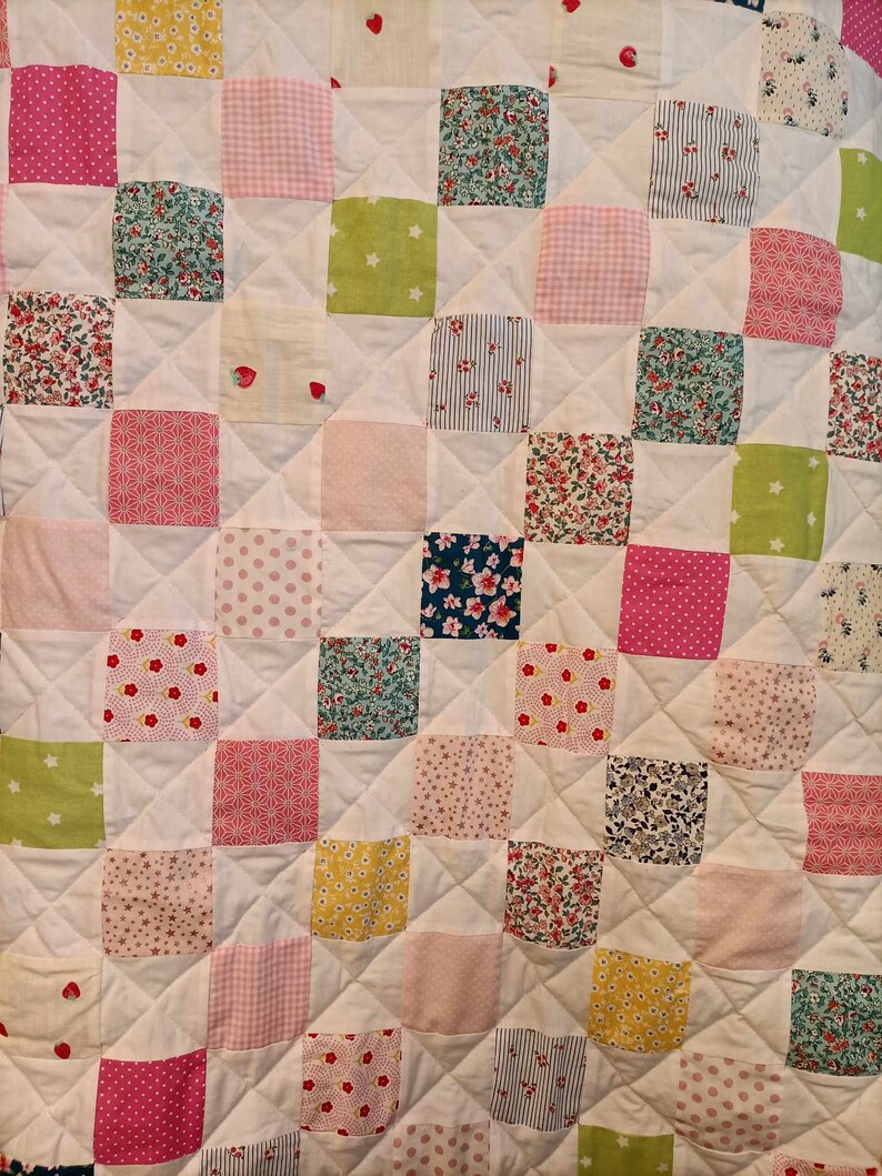 May include: A white quilt with a patchwork design. The quilt is made up of squares of different fabrics, each with a different pattern. The patterns include floral prints, polka dots, stripes, and geometric designs. The quilt is quilted in a diamond pattern.