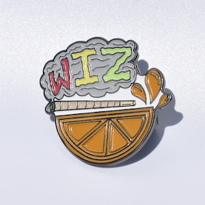May include: A colorful enamel pin featuring a cartoon orange slice with a lit joint and smoke forming the word "WIZ" in red, yellow, and green.