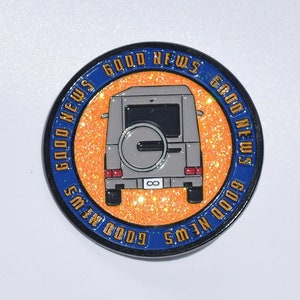 Peut inclure: Une épingle ronde en émail bleu et or avec les mots "Good News" répétés sur le bord. Le centre de l'épingle présente une voiture argentée sur un fond orange scintillant.