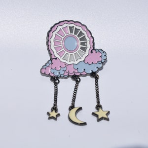 May include: A pink and blue enamel pin with a white and gray circle design, a crescent moon, and two stars hanging from chains.