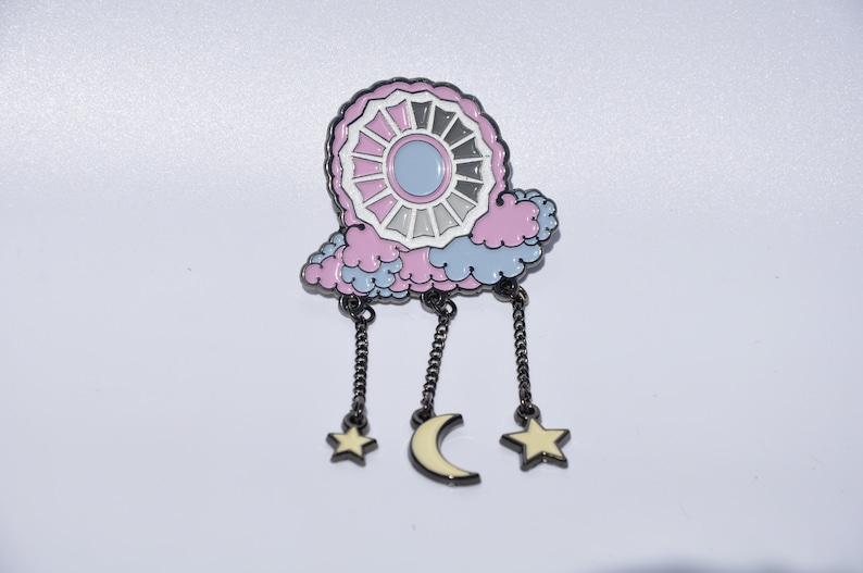 May include: A pink and blue enamel pin with a white and gray circle design, a crescent moon, and two stars hanging from chains.