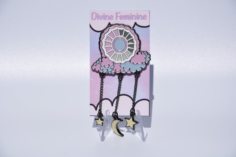 May include: A black enamel pin featuring a pink and blue cloud with a white and gray circle design inside. The cloud is hanging from three chains with a crescent moon and two stars at the bottom. The pin is on a white background with the text "Divine Feminine" above it.