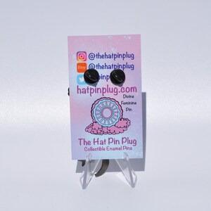 May include: A black enamel pin with a white design of a wheel with a pink and blue background. The pin is on a white card with the text "The Hat Pin Plug" and "@thehatpinplug" and "hatpinplug.com".