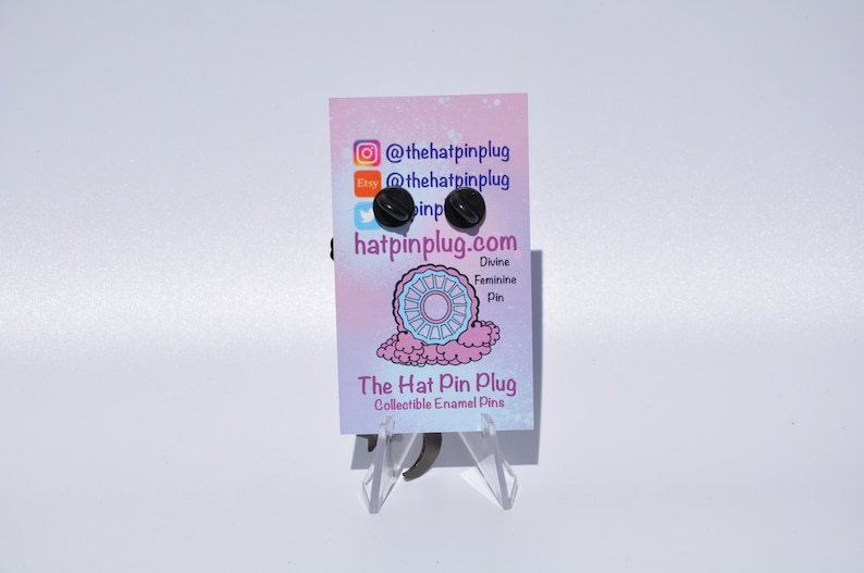 May include: A black enamel pin with a white design of a wheel with a pink and blue background. The pin is on a white card with the text "The Hat Pin Plug" and "@thehatpinplug" and "hatpinplug.com".