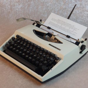 Può includere: Macchina da scrivere vintage color crema con tasti neri e carta inserita. La macchina da scrivere ha un design elegante con una leva di ritorno del carrello cromata e una copertura del nastro nera. La carta mostra del testo. La macchina da scrivere è su una superficie grigio chiaro.