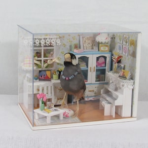 Puede incluir: Diorama de una habitación en miniatura con un pájaro que lleva un collar. La habitación cuenta con un escritorio con un portátil, un piano y una pequeña mesa con una taza de té. Las paredes están decoradas con papel pintado floral y obras de arte en miniatura.