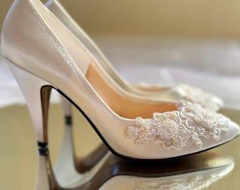 May include: A pair of ivory satin bridal shoes with a delicate lace and pearl embellishment on the toe.