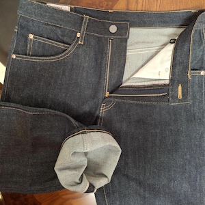 Pode incluir: Calças de ganga azul escuro com fecho de botão e zíper. As calças de ganga têm um design clássico de cinco bolsos com rebites de cobre e uma bainha dobrada, mostrando a borda da orla. As calças de ganga estão sobre uma superfície de madeira.