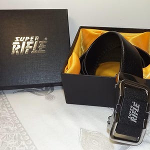 May include: A black leather belt with a silver buckle, presented in a black gift box lined with yellow satin. The box and buckle feature the text "SUPER RIFLE". The belt is a classic accessory.