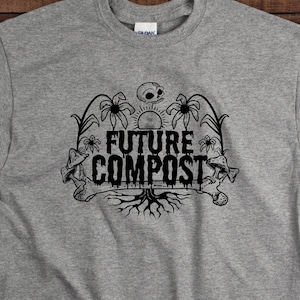 Puede incluir: Una camiseta gris con un diseño gráfico negro que presenta una calavera, hongos y el texto "FUTURE COMPOST".