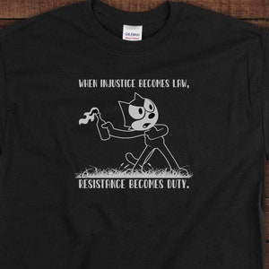 May include: Black t-shirt with a white graphic of a cartoon cat holding a Molotov cocktail. The text "When injustice becomes law, resistance becomes duty." is printed below the image.