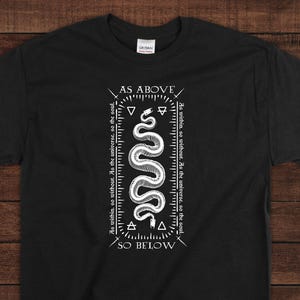 May include: Black t-shirt with a white graphic of a snake and the text "AS ABOVE" and "SO BELOW". The design includes geometric shapes and the phrase "As within, so without. As the universe, so the soul."