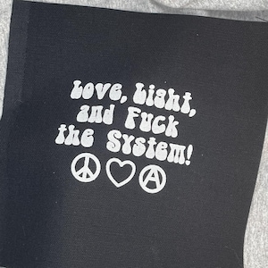May include: A square black fabric patch featuring white embroidered text that reads "Love, Light, and Fuck the System!" followed by a peace sign, a heart symbol, and an anarchy symbol. The embroidery has a bold, rounded, retro-style font.