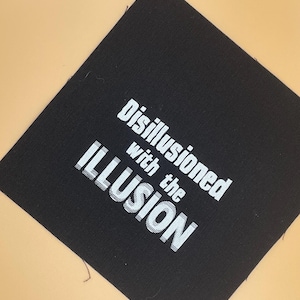 May include: A square black fabric patch featuring white embroidered text that reads Disillusioned with the ILLUSION. The lettering is arranged in three lines with the word ILLUSION appearing in a larger, bold, sans-serif font style.