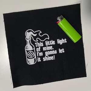 May include: Black fabric patch with white text and graphic of a bottle with flames, and the words "This little light of mine. I'm gonna let it shine!" A green lighter is next to the patch.