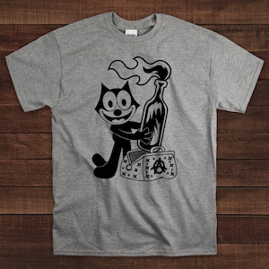 Puede incluir: Una camiseta gris con una ilustración en blanco y negro de un gato de dibujos animados sosteniendo un cóctel Molotov. El gato está de pie junto a una bolsa en blanco y negro con un símbolo de anarquía.