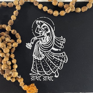 May include: A black canvas with a white illustration of a dancing figure, accompanied by text in a foreign language. A strand of wooden beads with a tassel hangs on the left side. The beads are a light brown color.