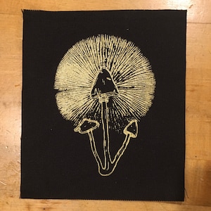 May include: A black fabric patch with a gold-colored illustration of a mushroom with a large cap and a stem with two smaller mushrooms branching off. The mushroom is stylized with a detailed design.