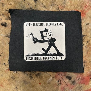 May include: Black and white patch featuring a cartoon cat holding a Molotov cocktail. The text reads "When injustice becomes law, resistance becomes duty."
