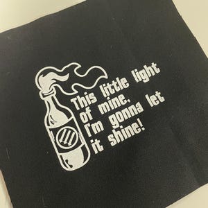 Peut inclure: Carré de tissu noir avec texte blanc et une illustration de bouteille. Le texte dit : « This little light of mine, I'm gonna let it shine ! » La bouteille a une flamme qui sort du haut.