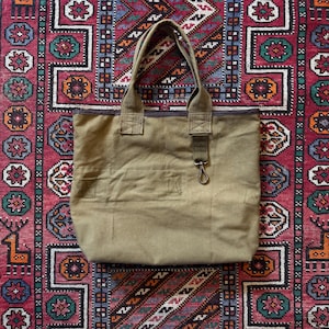 May include: A khaki canvas tote bag with strong handles and a zip closure. The bag has a small front pocket and a metal clasp. The bag is placed on a patterned red, blue, and green rug.