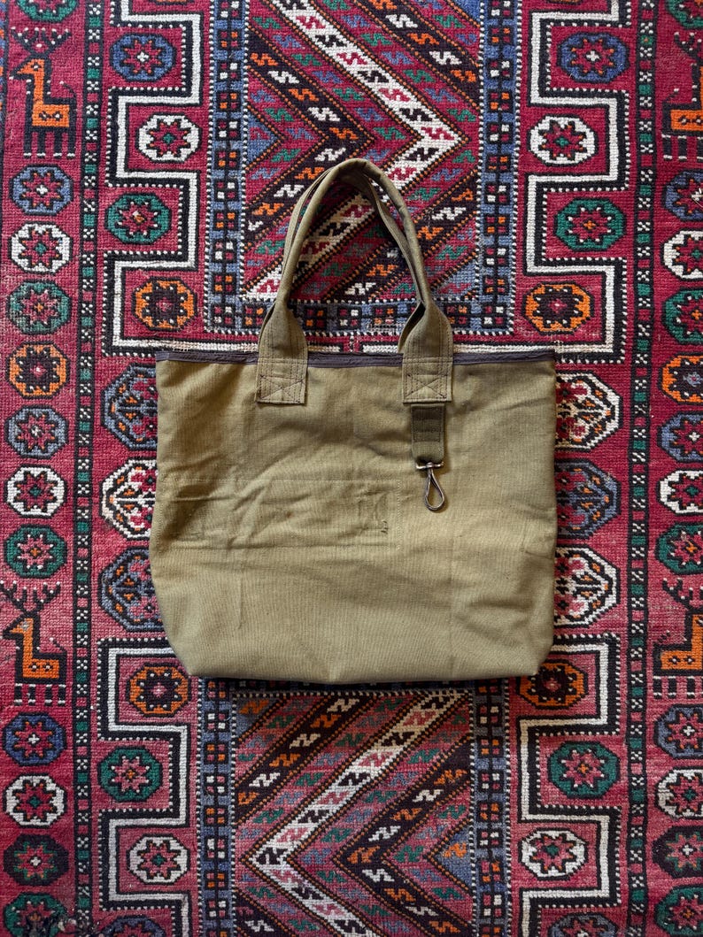 May include: A khaki canvas tote bag with strong handles and a zip closure. The bag has a small front pocket and a metal clasp. The bag is placed on a patterned red, blue, and green rug.