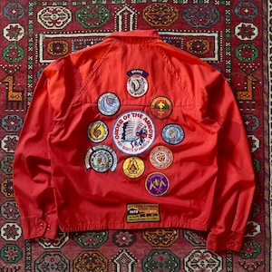 May include: A red vintage jacket adorned with numerous patches, including "Order of the Arrow" and "Boy Scouts of America" badges. The jacket features a button-up design and a collar. The patches display diverse colours, shapes, text, and graphics.