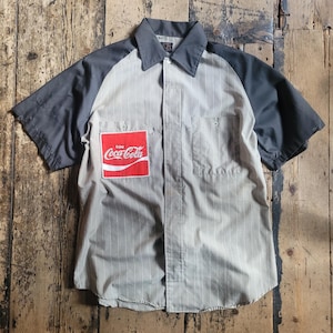 May include: A short-sleeved work shirt with a grey collar and sleeves, and a light beige body with vertical stripes. A red Coca-Cola logo patch is on the chest. The shirt has two chest pockets and a button-down front.