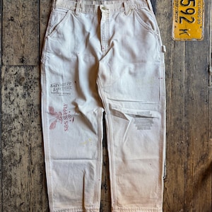 Könnte beinhalten: Cremefarbene Arbeitshose mit mehreren Taschen und einem Knopfverschluss. Die Hose hat Farbspritzer und verblasste Bereiche, mit Text und einer Grafik auf dem linken Bein. Ein Vintage-Kennzeichen aus Illinois ist im Hintergrund.