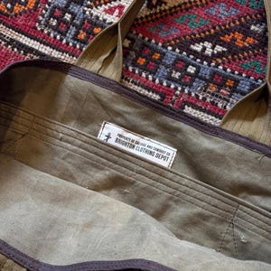 May include: An olive green canvas tote bag with brown trim and straps. The interior has a label reading "PROPERTY OF SALVAGE AND SAWDUST CO BRIGHTON CLOTHING DEPOT". The bag is open, showing the inside.