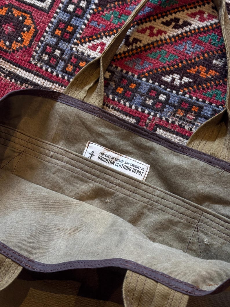 May include: An olive green canvas tote bag with brown trim and straps. The interior has a label reading "PROPERTY OF SALVAGE AND SAWDUST CO BRIGHTON CLOTHING DEPOT". The bag is open, showing the inside.