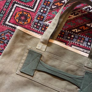May include: Olive green canvas tote bag with a reinforced handle and a contrasting green accent. The bag has a rectangular shape and a sturdy design, suitable for carrying various items. The background features a colorful patterned rug.