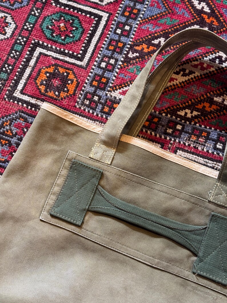 May include: Olive green canvas tote bag with a reinforced handle and a contrasting green accent. The bag has a rectangular shape and a sturdy design, suitable for carrying various items. The background features a colorful patterned rug.