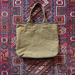 May include: A khaki canvas tote bag with strong handles. The bag is rectangular and simply designed. The background is a colourful, patterned rug with red, blue, and white geometric designs.
