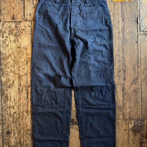 Puede incluir: Pantalones de trabajo azul oscuro con bragueta de botones, bolsillos delanteros y corte relajado. Los pantalones están hechos de una tela duradera y tienen un diseño clásico y utilitario. Los pantalones se muestran sobre una superficie de madera.