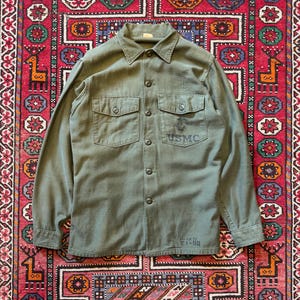 Può includere: Camicia verde oliva in stile militare con due tasche sul petto e le lettere "USMC" stampate sul petto destro. La camicia ha maniche lunghe e un colletto. Il testo "VALER TE 81-88" è stampato in basso a destra.