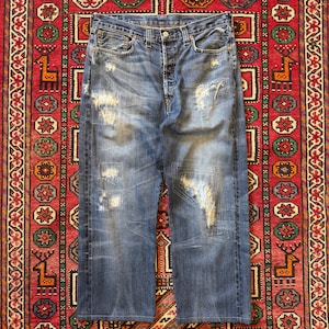 Op de afbeelding: Verbleekte blauwe denim jeans met distressed details, waaronder scheuren en patches. De jeans hebben een klassiek ontwerp met vijf zakken en een knoopsluiting. De jeans worden gepresenteerd op een patroonkleed.