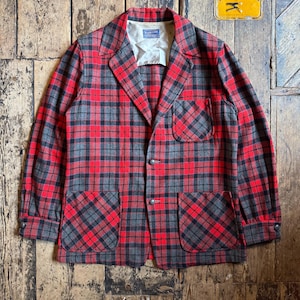 May include: A vintage red, black, and grey plaid blazer with two front pockets and two buttons. The jacket is laid flat on a wooden surface. The blazer has a classic, tailored design, and a cream-coloured lining is visible at the collar.