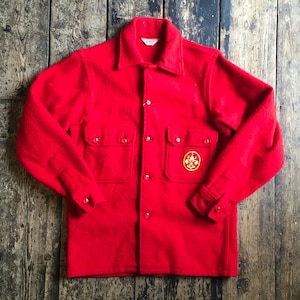 Puede incluir: Una chaqueta de lana roja con un parche en el pecho izquierdo que presenta un diseño de flor de lis amarillo. La chaqueta tiene dos bolsillos grandes en la parte delantera y un cierre de botones.
