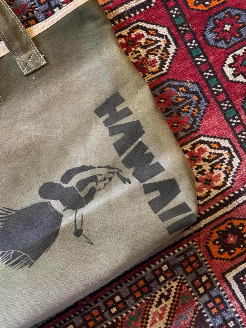 May include: Olive green tote bag with the word "HAWAII" printed in black block letters. A black silhouette of a hula dancer is also printed on the bag. The bag has a white trim and a canvas handle.