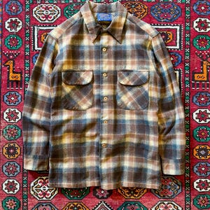 Puede incluir: Una camisa abotonada a cuadros marrón, beige y azul con dos bolsillos en el pecho. La camisa tiene cuello y está hecha de una tela suave y texturizada. La camisa se muestra sobre una alfombra roja estampada.