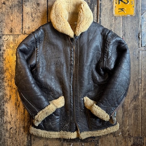 Puede incluir: Chaqueta bomber de cuero marrón con cuello y forro de borrego beige. La chaqueta tiene cremallera, dos bolsillos delanteros y ribete de borrego en los puños y el dobladillo. Sobre una superficie de madera.