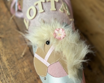 May include: A pink and brown school cone with a horse head decoration. The horse has a white mane and tail, a pink bridle, and a black eye. The cone is decorated with a pink ribbon, a white flower, and the name "OTTA" in white letters.