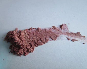 May include: A close-up of a sample of loose, shimmery, rose gold mica powder. The powder is spread out on a white surface, showing its texture and color.
