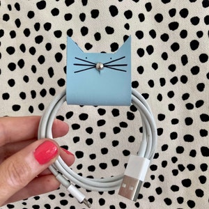 May include: A pale blue cat-shaped cable tidy with black whiskers and a silver stud. It holds a white cable with a USB connector. The background is a white fabric with black polka dots.