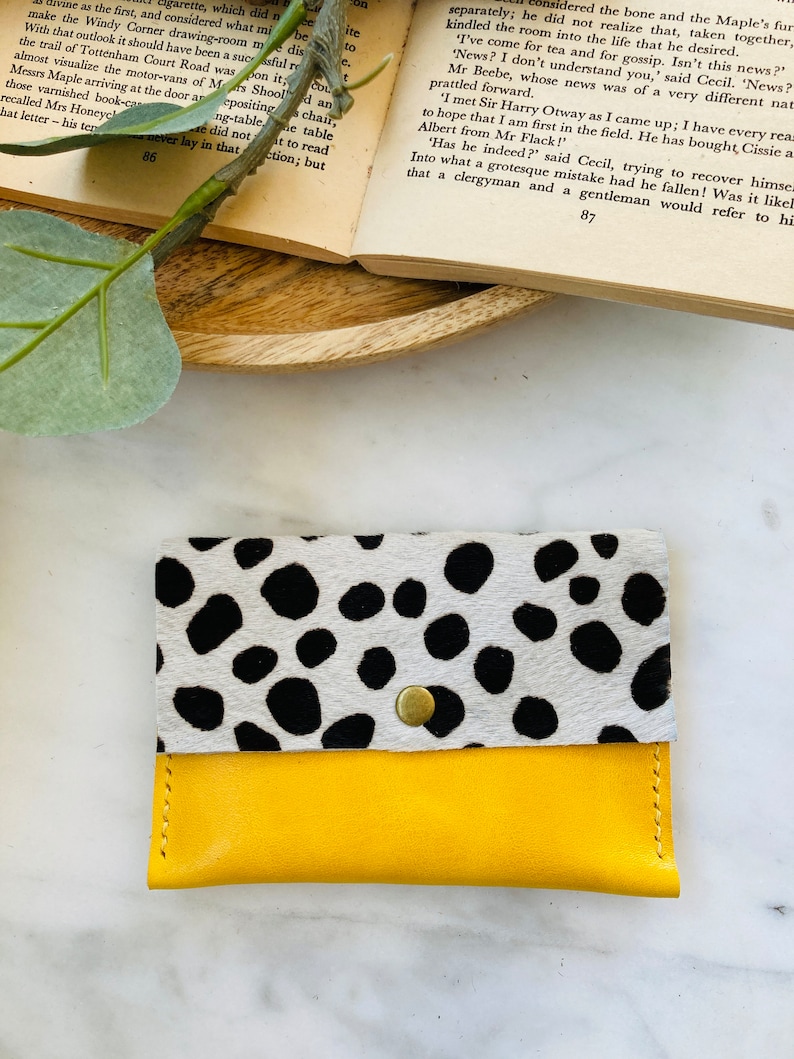 May include: A handmade cardholder wallet with a unique design. The top flap features a cowhide pattern with black spots on a white background. The bottom section is bright yellow leather, secured with a brass snap.