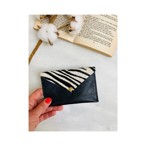 May include: Black leather cardholder with a zebra print flap secured by a gold-colored snap. The cardholder is rectangular and has a folded flap. The zebra print is black and white.