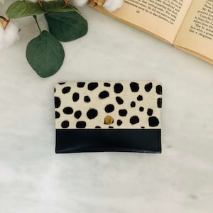 May include: A small, rectangular cardholder with a black leather bottom and a top section featuring a cream-colored, spotted pattern. A gold-colored snap closure is centered on the front. The cardholder is likely made of leather.