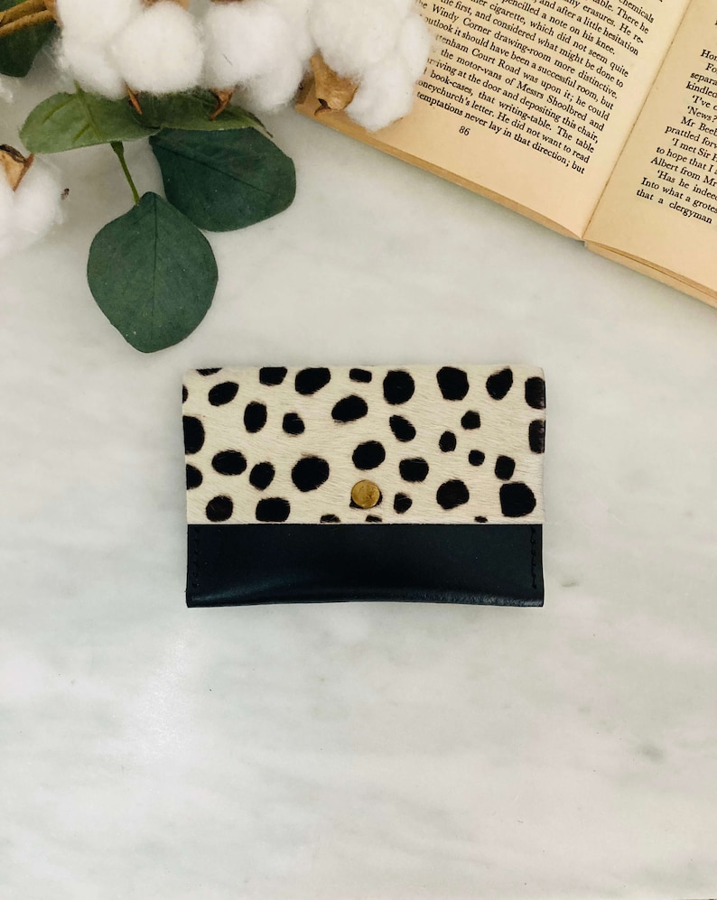 May include: A small, rectangular cardholder with a black leather bottom and a top section featuring a cream-colored, spotted pattern. A gold-colored snap closure is centered on the front. The cardholder is likely made of leather.